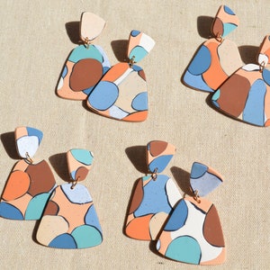 Könnte beinhalten: Ein Set mit sechs bunten, abstrakten, geometrischen Ohrringen. Jeder Ohrring besteht aus zwei Teilen aus Ton, wobei ein Teil größer ist als der andere und ein Muster in Blau, Orange, Braun und Weiß aufweist. Die Ohrringe sind an goldenen Haken befestigt.