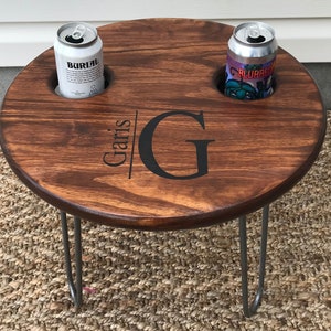 May include: A round wooden table with two drink holders and a personalized inscription that reads "Garis". The table has a dark brown wood finish and is supported by two hairpin legs.