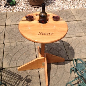 May include: A round, light-colored wooden wine table with a bottle of wine and two glasses. The table has a central stem and a three-legged base. The word "Stone" is engraved on the table's surface.
