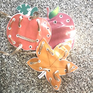 May include: A set of three fall-themed lacing cards featuring a pumpkin, apple, and leaf. Each card has holes around the edges for threading a ribbon or string.