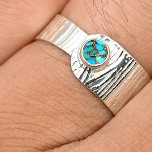 May include: A silver ring featuring a round turquoise gemstone. The ring has a textured band with a distinctive pattern. The turquoise stone is a vivid blue-green hue, set in a circular bezel.
