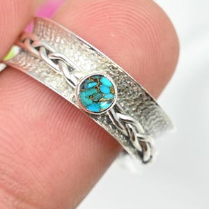 May include: A silver ring featuring a turquoise gemstone. The ring has a textured band with a braided pattern and a small, round turquoise stone at the centre. The ring is held by a person's fingers.