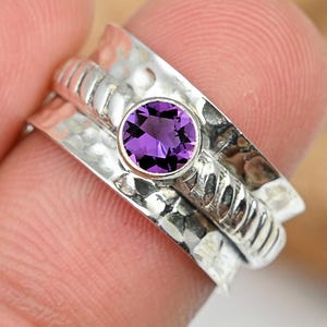 May include: A silver ring with a hammered texture and a spinning band. The ring features a round, vibrant purple gemstone at its centre. The ring is shown close up, highlighting the details of the metalwork and the gemstone.