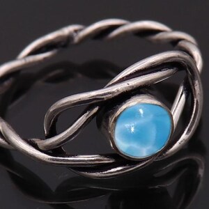 Könnte beinhalten: Ein silberner Ring mit einem geflochtenen Banddesign. Der Ring hat einen runden, hellblauen Edelstein in der Mitte. Der Ring wird auf einer dunklen Oberfläche präsentiert, die seine komplizierten Details und Farbe hervorhebt.