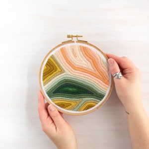 May include: A circular embroidery hoop with a colorful, abstract design. The design features a variety of shades of green, yellow, orange, and pink, creating a layered, textured effect.