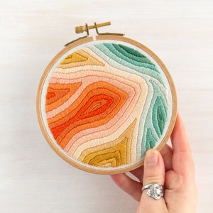 May include: A circular embroidery hoop with a colorful abstract design. The design features a variety of shades of orange, yellow, pink, and green, creating a layered and textured effect.