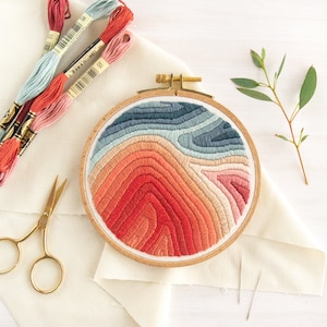 May include: A close-up of a circular embroidery hoop with a colorful, abstract design. The design features a gradient of red, orange, and blue hues, creating a layered, wave-like pattern. The hoop is made of wood and has a natural finish.
