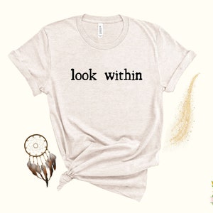 May include: A light beige t-shirt with the text "look within" printed in black on the front. The shirt is made of a soft, comfortable material and has a relaxed fit.