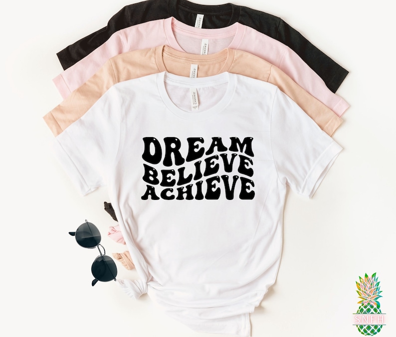 May include: White t-shirt with black text that reads "Dream Believe Achieve" in a retro font style. The shirt is folded and stacked on top of other t-shirts in pink and black.
