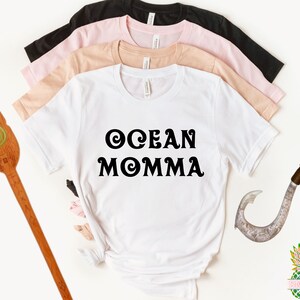 May include: A white t-shirt with the text "OCEAN MOMMA" in black, surrounded by other t-shirts in black, pink, and tan. Decorative wooden paddle and fish hook props are on the sides. A pineapple logo is in the bottom right corner.