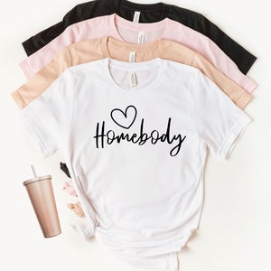 May include: White t-shirt with a black heart and the text "Homebody" in black script font. The shirt is shown folded on top of a stack of three other t-shirts in black, pink, and beige.