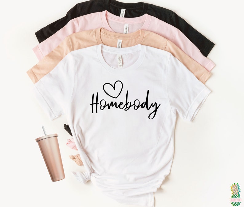 May include: White t-shirt with a black heart and the text "Homebody" in black script font. The shirt is shown folded on top of a stack of three other t-shirts in black, pink, and beige.