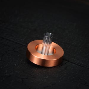 May include: A copper-colored metal spinning top with a silver-colored central post. The top has a circular base and a cylindrical post. The object is set against a dark, textured background.