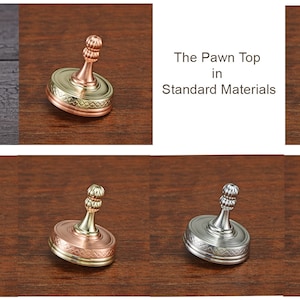 May include: A collection of metal spinning tops in various color combinations. The tops have a rounded base with a decorative pattern and a textured stem. The text "The Pawn Top in Standard Materials" is also visible.