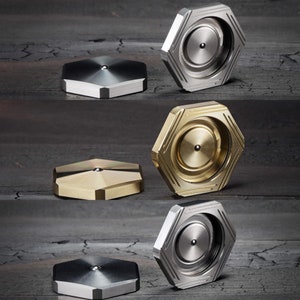 May include: Four hexagonal metal spinning tops in different colors and finishes. The tops are made of brass, stainless steel, and titanium. They are all designed with a central bearing and a smooth, rounded edge.