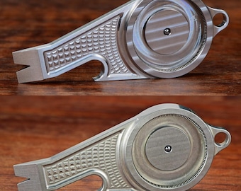May include: A silver metal bottle opener with a circular design and a textured surface. The opener has a small hole on the side and a bottle opener on the bottom.