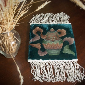May include: A green velvet cushion with a white fringe border. The cushion features an embroidered design of a vase with a floral pattern.