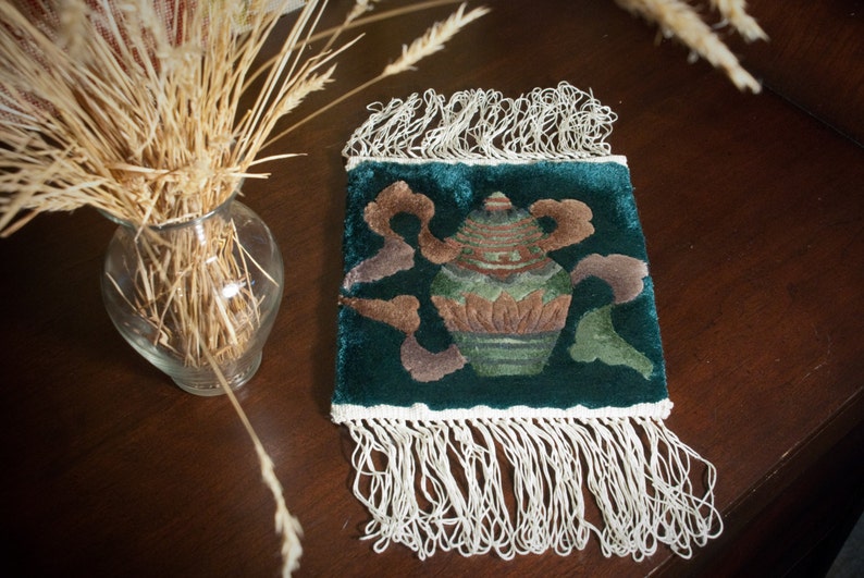May include: A green velvet cushion with a white fringe border. The cushion features an embroidered design of a vase with a floral pattern.