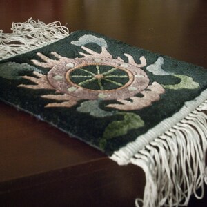 May include: A small, rectangular rug with a dark green background and a central design featuring a pink and green floral pattern. The rug has white fringe along the edges, adding a decorative touch. The rug is placed on a dark brown surface.