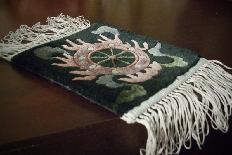 May include: A small, rectangular rug with a dark green background and a central design featuring a pink and green floral pattern. The rug has white fringe along the edges, adding a decorative touch. The rug is placed on a dark brown surface.