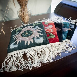 May include: A collection of colorful, embroidered textile squares with fringe. The squares feature intricate designs in shades of green, red, black, and blue. They are arranged on a dark brown surface, with a vase of wheat stalks in the background.
