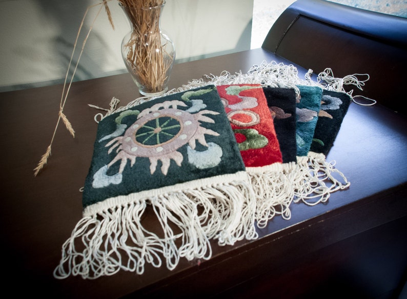 May include: A collection of colorful, embroidered textile squares with fringe. The squares feature intricate designs in shades of green, red, black, and blue. They are arranged on a dark brown surface, with a vase of wheat stalks in the background.