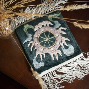 May include: A dark teal rectangular textile with an embroidered design featuring a central wheel-like motif in pink and green, surrounded by stylized floral elements. The textile has a white fringe along the bottom edge.