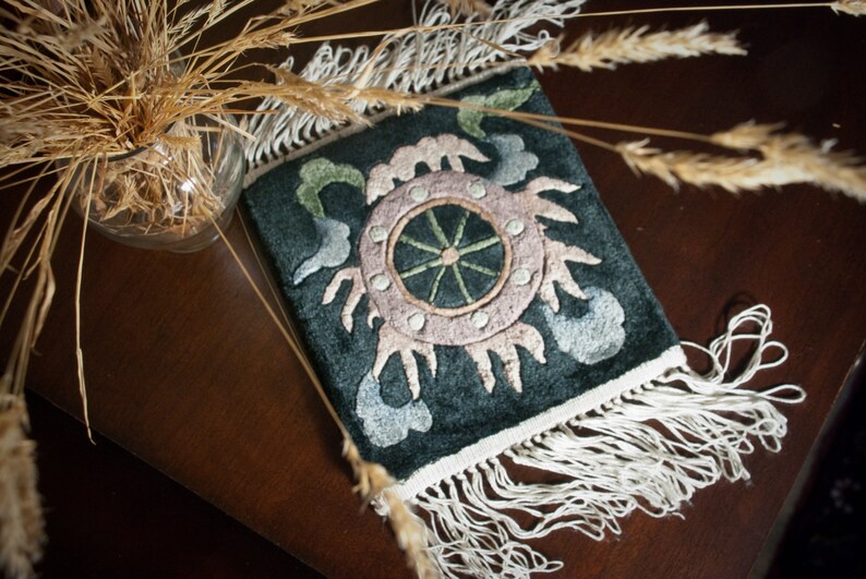 May include: A dark teal rectangular textile with an embroidered design featuring a central wheel-like motif in pink and green, surrounded by stylized floral elements. The textile has a white fringe along the bottom edge.