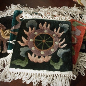 May include: A collection of square, dark-colored decorative rugs with intricate, colorful designs. Each rug features a unique pattern, including a horse, a sun-like emblem, and floral motifs. The rugs have white fringe along the edges, adding a textured border.