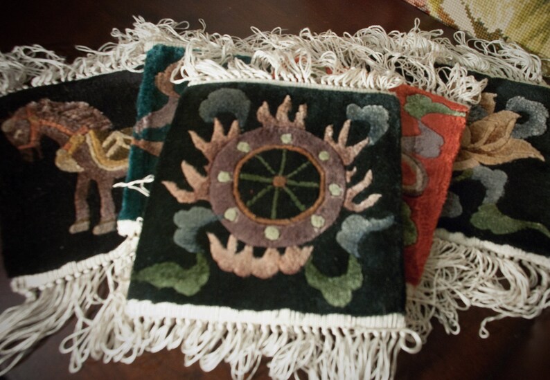 May include: A collection of square, dark-colored decorative rugs with intricate, colorful designs. Each rug features a unique pattern, including a horse, a sun-like emblem, and floral motifs. The rugs have white fringe along the edges, adding a textured border.