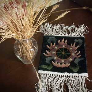 May include: A decorative, dark green textile with a sun-like design in brown and green, featuring a white fringe. A clear glass vase holds dried wheat stalks, set on a dark wooden surface.