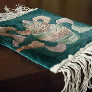 May include: A green and white fringed throw rug with a floral design. The rug has a white fringe border and a floral pattern in shades of green, white, and brown.