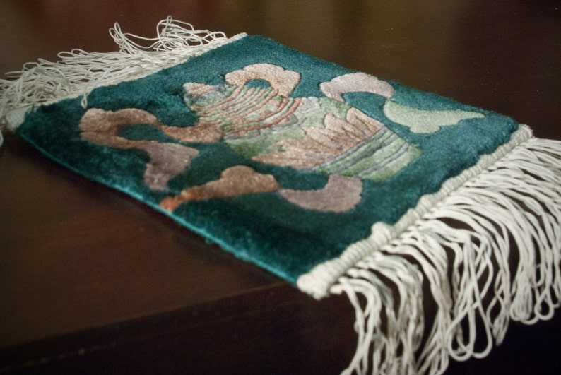 May include: A green and white fringed throw rug with a floral design. The rug has a white fringe border and a floral pattern in shades of green, white, and brown.