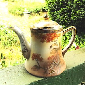 May include: Antique porcelain coffee pot with a lid, featuring a floral design in shades of orange, brown, and gold. The pot has a curved spout, a handle, and gold trim. The background is a blurred outdoor scene.