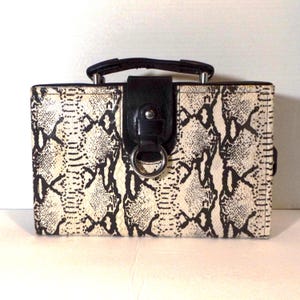 May include: A rectangular handbag with a snakeskin pattern in black and cream. The bag features a black leather handle and a black leather flap with a silver ring closure. The bag is designed with a structured shape.
