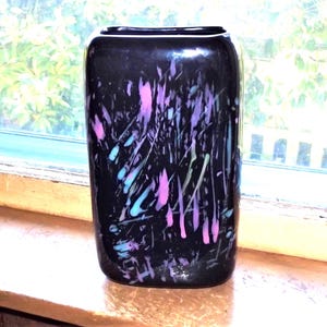 May include: A rectangular glass vase with a glossy black base color, featuring an abstract splatter pattern of vibrant pink and light blue paint-like streaks across the front surface, with rounded corners and a wide, open top opening.