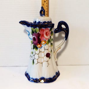 May include: A white porcelain teapot with a blue and gold trim. The teapot is decorated with a floral pattern of pink, purple, and yellow roses. The teapot has a lid with a knob on top.
