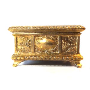 May include: A rectangular, gold-colored jewelry box with ornate detailing. The box features a hinged lid and is raised on four small feet. The front of the box has a central oval design flanked by floral patterns. The box is made of a shiny, metallic material.