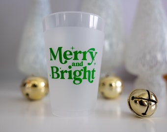 May include: A frosted plastic cup with the words "Merry and Bright" in green lettering. The cup is sitting on a white surface with gold jingle bells around it.