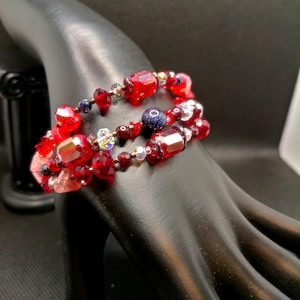 May include: A multi-strand bracelet featuring a variety of red faceted glass beads, dark blue glittery beads, and clear iridescent accents. The beads are strung on a flexible wire, creating a layered look with different shapes and sizes of beads.