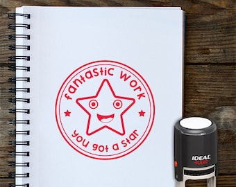 May include: Red and white teacher's stamp with a star and the text "Fantastic Work You Got A Star". The stamp is on a white notebook with a spiral binding. The stamp is on a wooden surface.