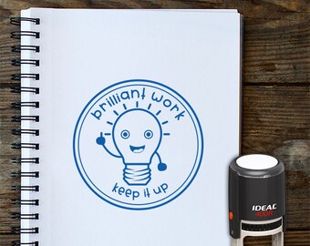 May include: Blue and white teacher stamp with a lightbulb graphic and the text "Brilliant Work Keep It Up". The stamp is on a white notebook with a spiral binding. The text "Teachers Stamp" is printed below the stamp.