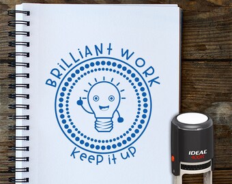 May include: A blue and white teacher stamp with the text "Brilliant Work Keep it Up" and a cartoon lightbulb with a smiling face. The stamp is on a white notebook with a black spiral binding. The text "Teachers Stamp" is printed below the stamp.