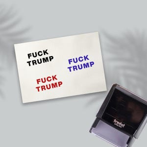 May include: A white paper with the words "FUCK TRUMP" stamped in black, blue, and red ink. A black and gray stamp is in the lower right corner, with the brand name "trodat" visible.