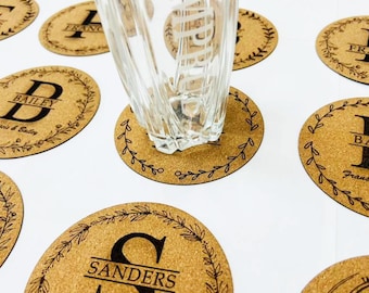 May include: A clear glass sits on a cork coaster with a black leaf design. Several oval cork coasters are visible, each with a different initial and last name in a black font. The coasters are on a white surface.