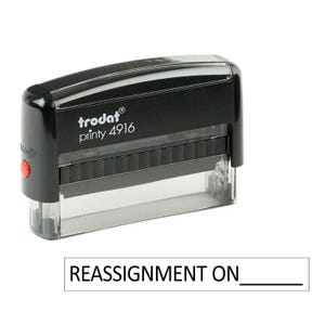 May include: Black self-inking stamp with the text "trodat printy 4916" and a red button. The stamp is designed to create a custom imprint that reads "REASSIGNMENT ON ______".