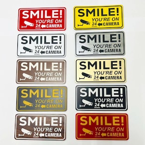 May include: Rectangular signs with the text "SMILE! YOU'RE ON 24 HOURS CAMERA" in various colors, including red, yellow, silver, brown, and black. Each sign features a security camera graphic, suitable for home or business security.