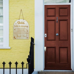 May include: A wooden sign with the text "Shhh... baby is sleeping. Please do not knock or ring doorbell. Please call or text. Packages can be left at the door. Thank you!" hanging on a yellow wall next to a brown wooden door.