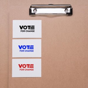 May include: Three white cards with the text "VOTE FOR CHANGE" in black, blue, and red. The cards are on a brown surface, held by a silver clipboard.
