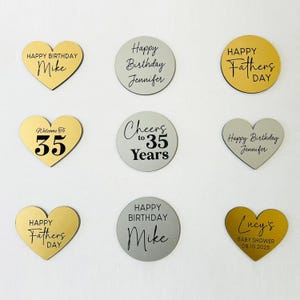 May include: A collection of gold and silver-colored decorative pieces with various celebratory messages. Heart-shaped and circular designs feature phrases like "Happy Birthday," "Happy Father's Day," and "Baby Shower." Some pieces include names and ages.